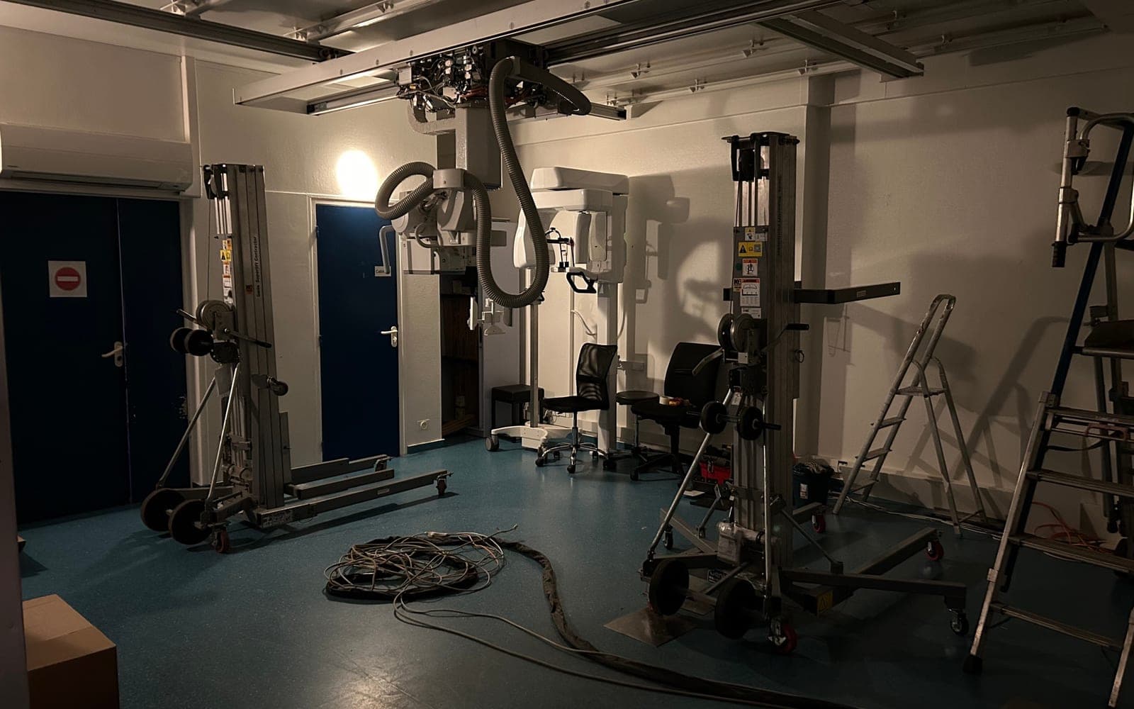 Salle de radiographie en cours de démontage : tube plafonnier décroché, monte-charges positionnés, câbles au sol