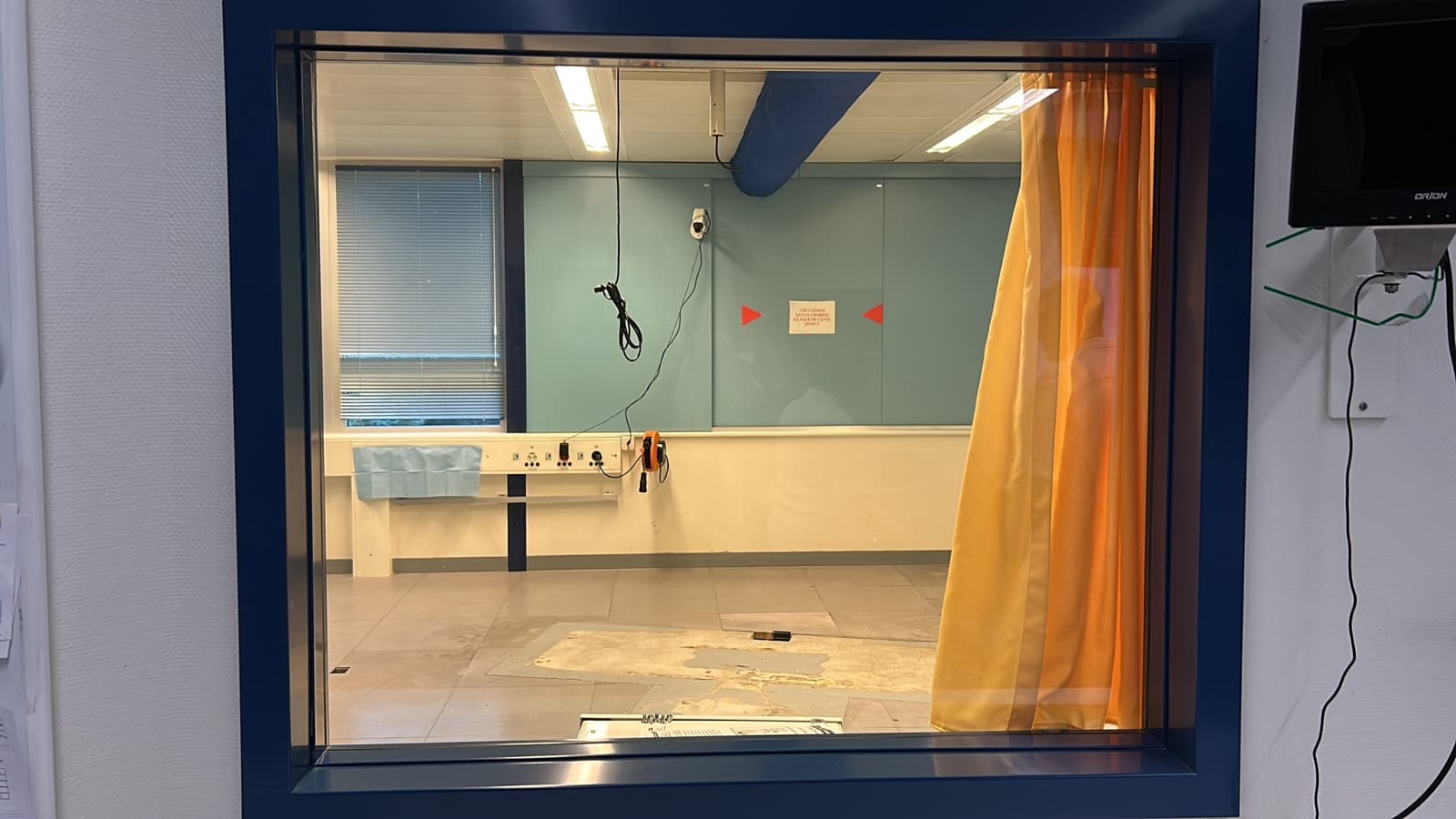 Salle de radiologie vidée vue à travers la vitre plombée, traces au sol où était le gantry
